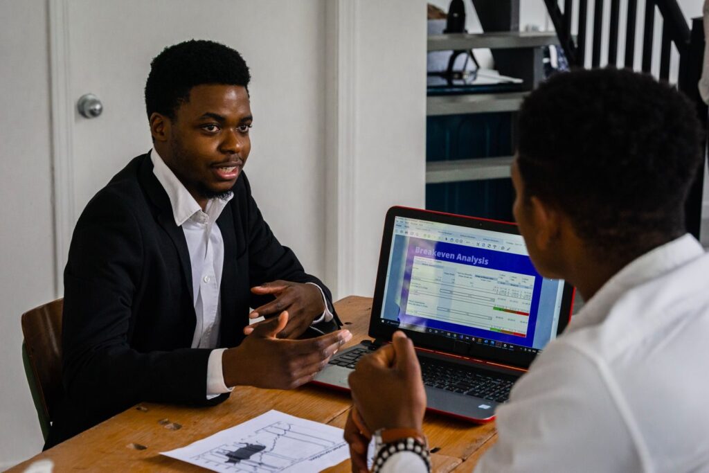 Two businessmen engage in a conversation about financial strategies with a laptop displaying a breakeven analysis.